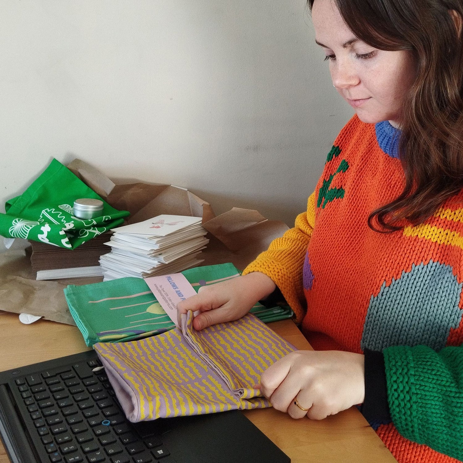 Ruth is packing an order of cushion covers on the table. She is wearing an orange jumper with colourful shapes on it.